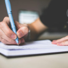 Persona sosteniendo bolígrafo azul escribiendo en el cuaderno.