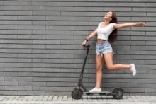 Una mujer posa felizmente parada en una pierna sobre un monopatín eléctrico